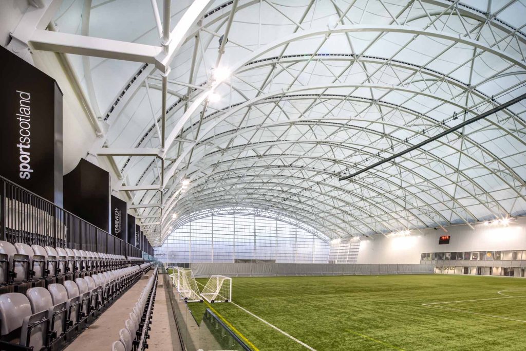 Oriam Sports Performance Centre interior showing curved steel roof structure inspired by Roberto Carlos free kick trajectory with football pitch and spectator seating