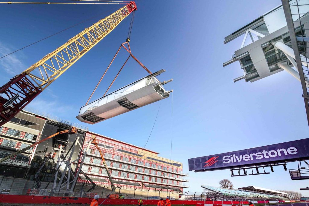 Silverstone Circuit bridge installation showing massive steel section being lifted by crane over Formula One racing track with Silverstone branding visible